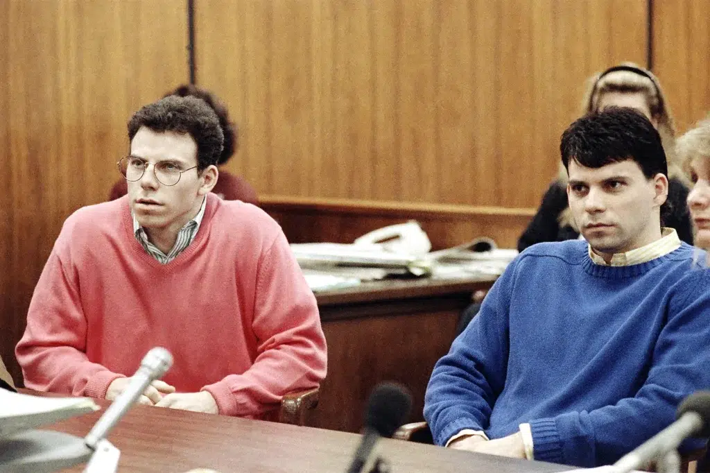 Two individuals seated in a courtroom setting, one wearing a red sweater, the other in blue. Menendez Brothers Case