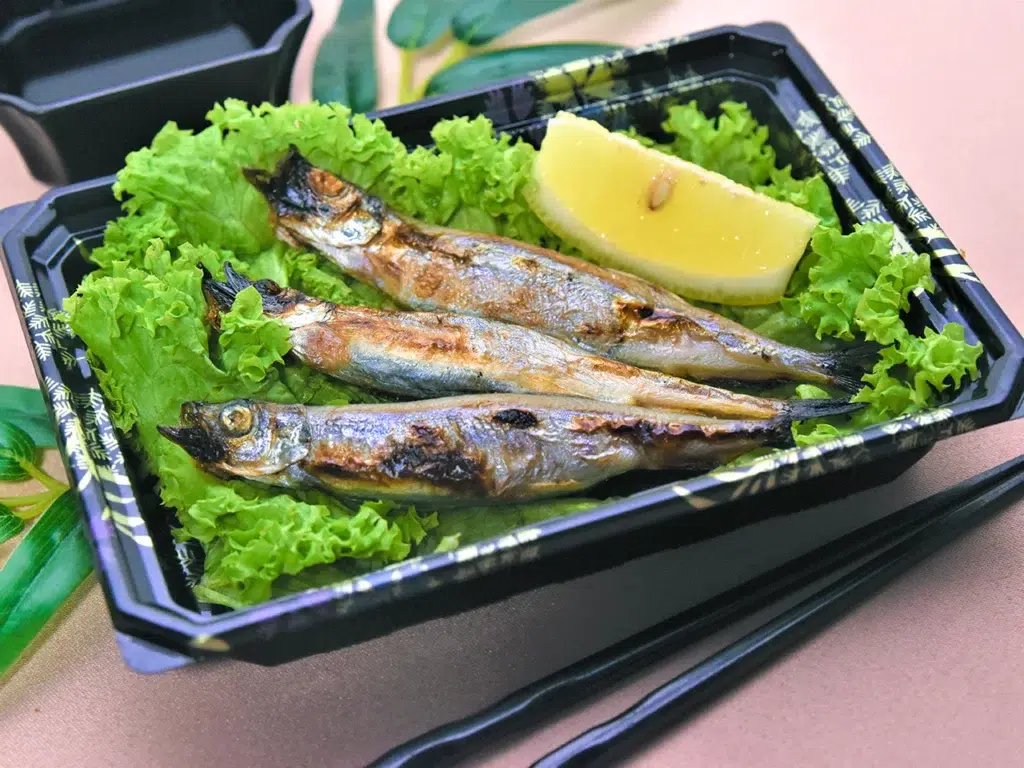 Grilled sardines with lettuce and lemon slice in a black takeout container. Shishamo