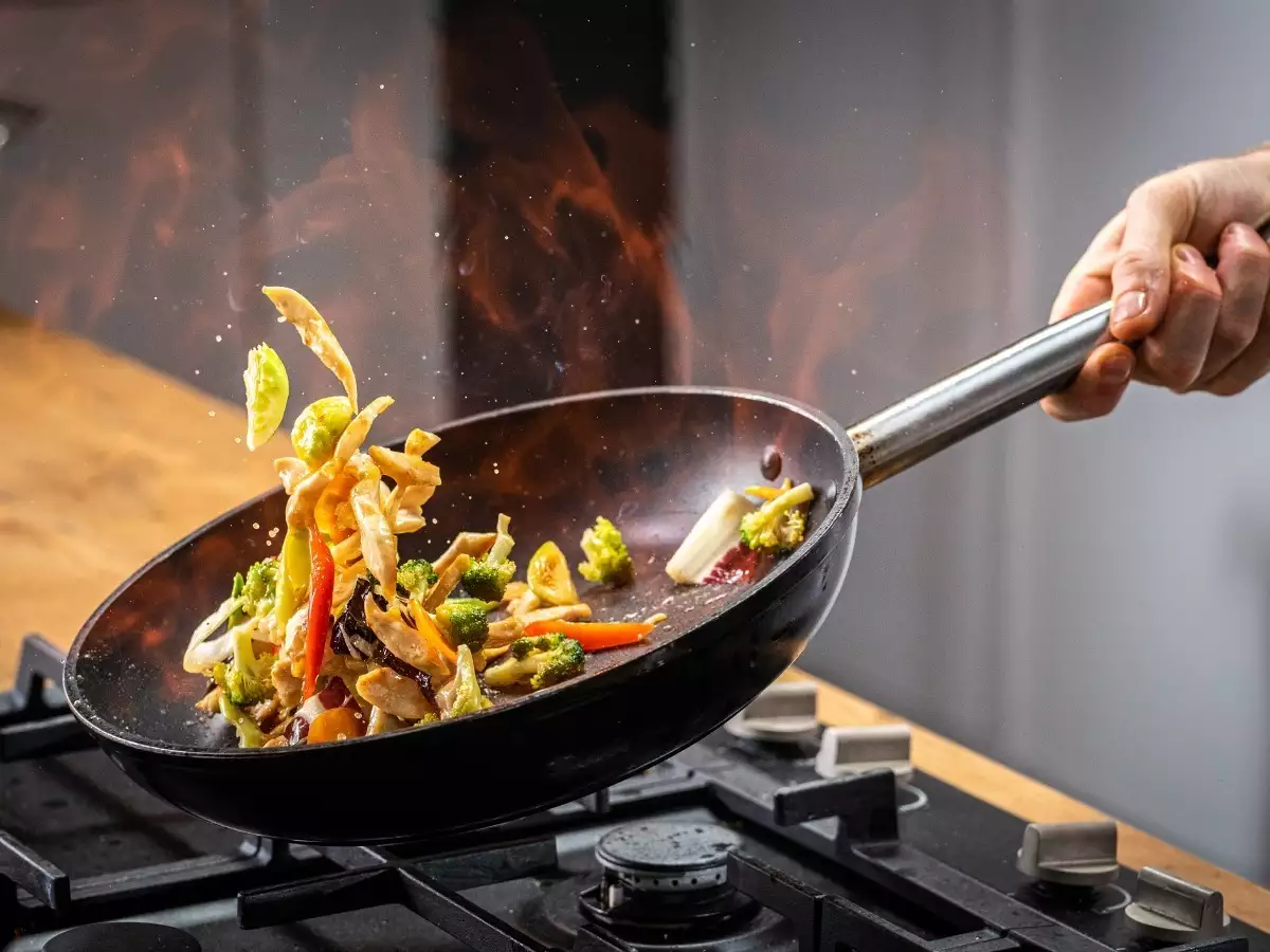 Vegetables being stir-fried in a wok with flames, hand holding the pan handle. Stir frying