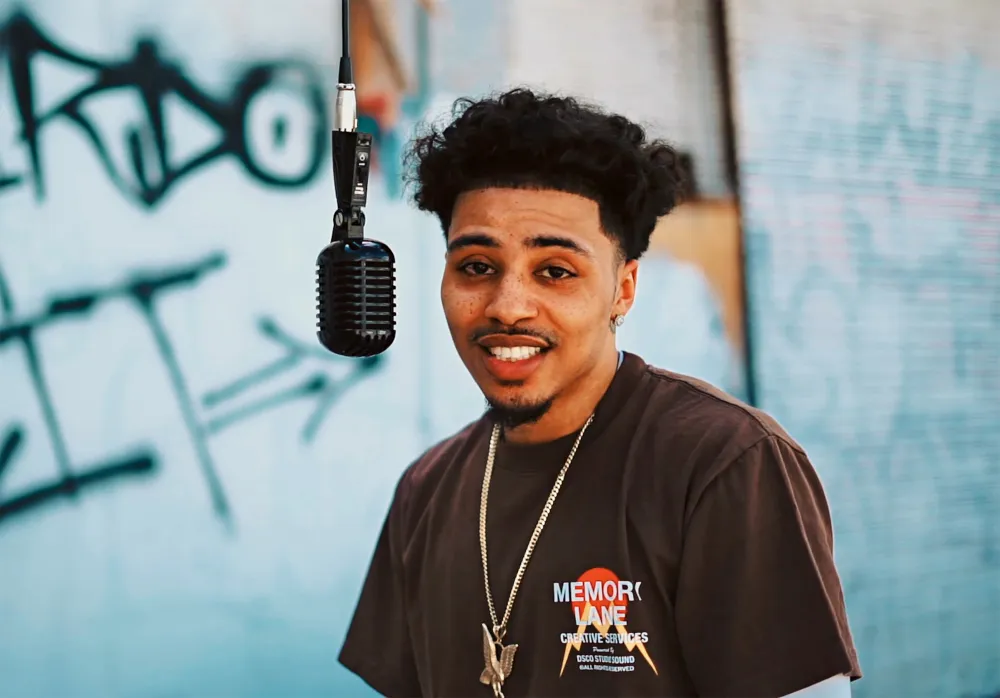 Person with microphone wearing a t-shirt and necklace, standing in front of a graffiti wall. Lucas Coly death